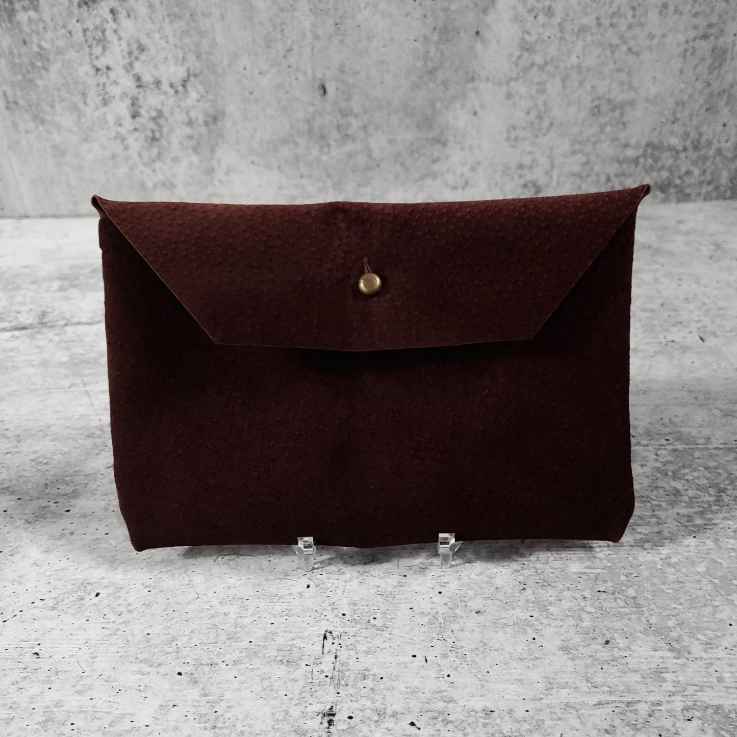 Front facing view of "soft suede clutch trapezoid" in chocolate against a concrete background.