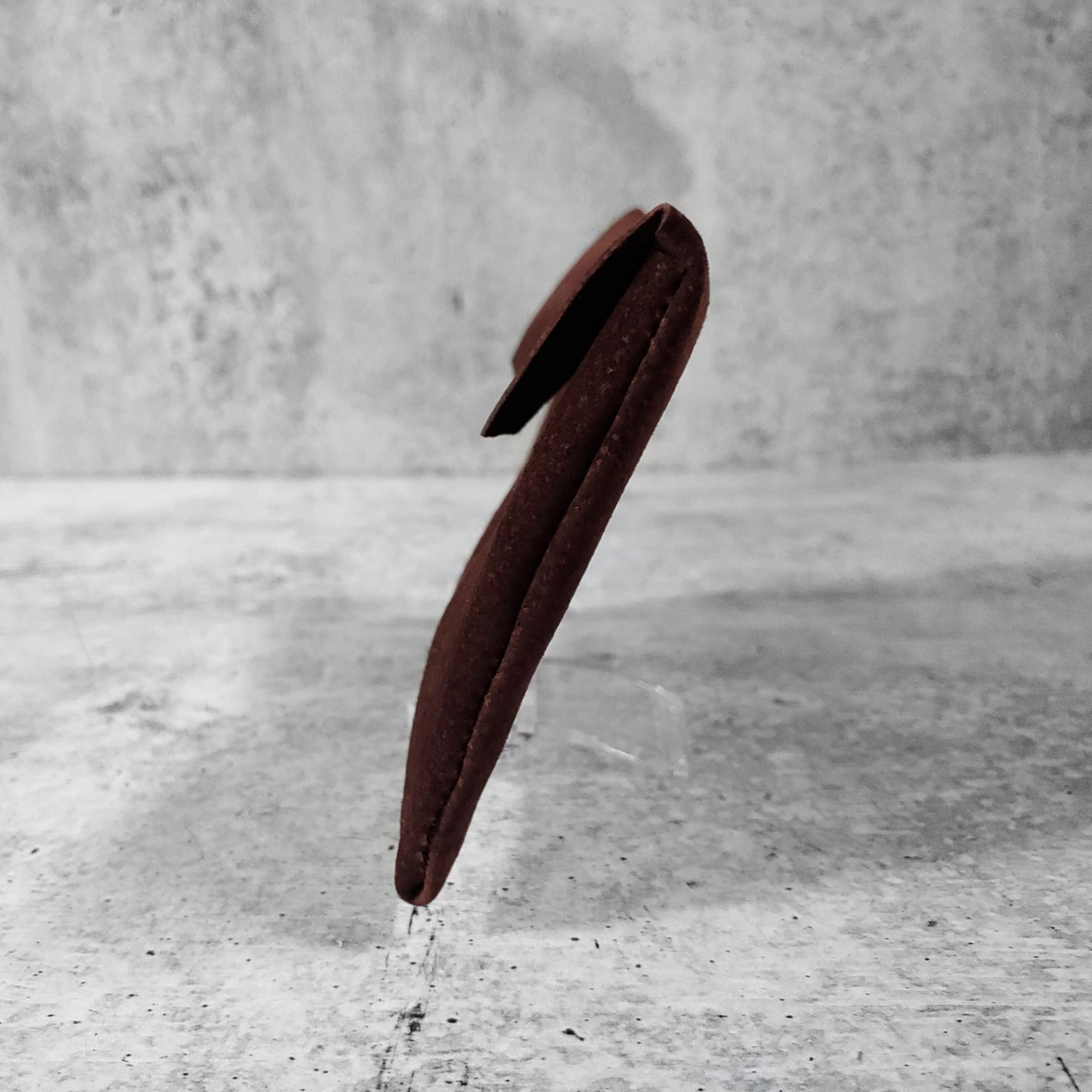 Side view of "soft suede clutch trapezoid" in chocolate against a concrete background.