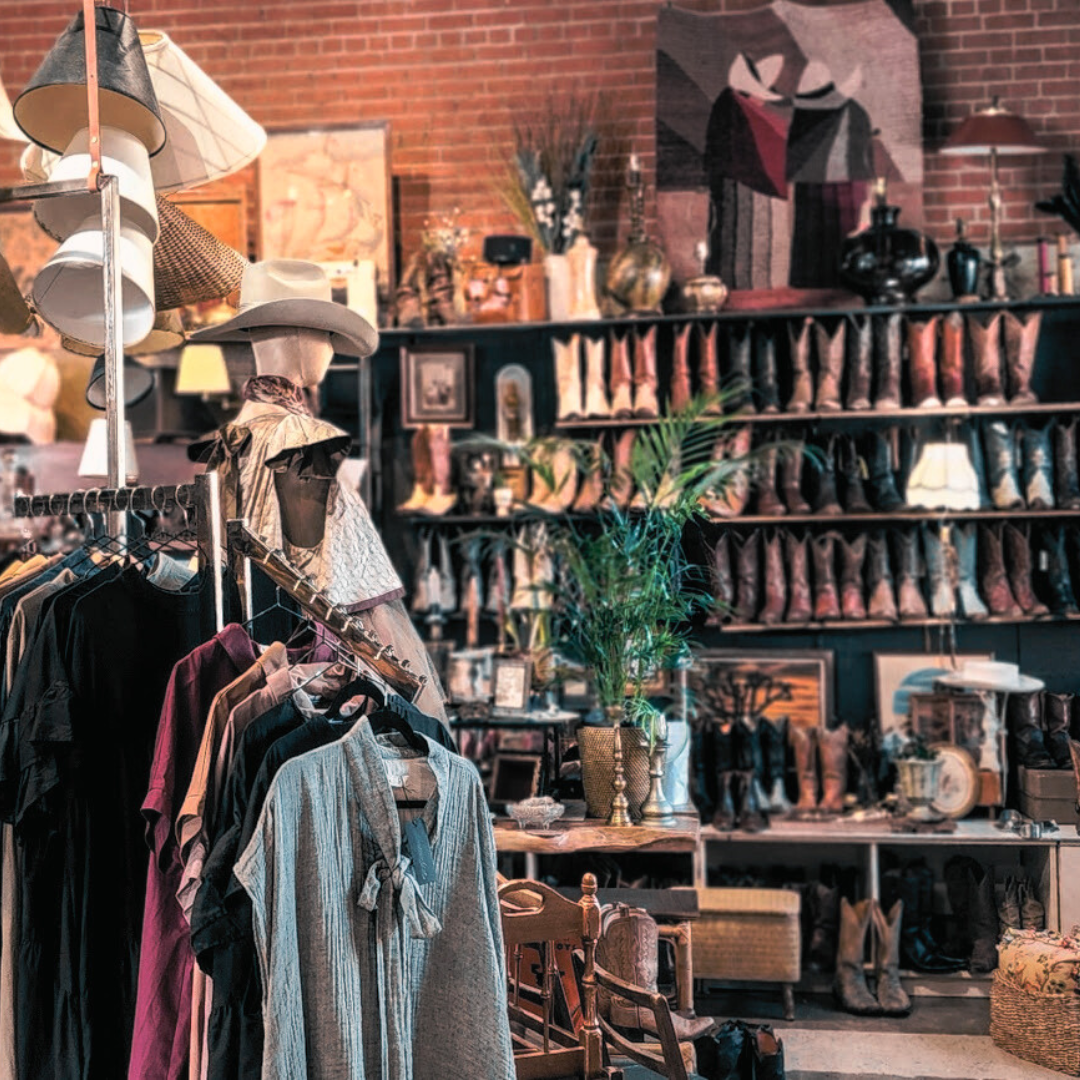 Selection of leather cowboy boots against a brick wall with McCULLOUGH clothing on display to the left. Mannequin wears a cowboy hat, blouse and tulle skirt. Lampshades hang from the ceiling and vintage home decor items fill the rest of the frame.