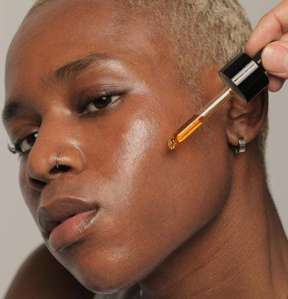 Amber colored NOTO Mini Deep Serum product being applied to African American model using the drop pipette applicator top.